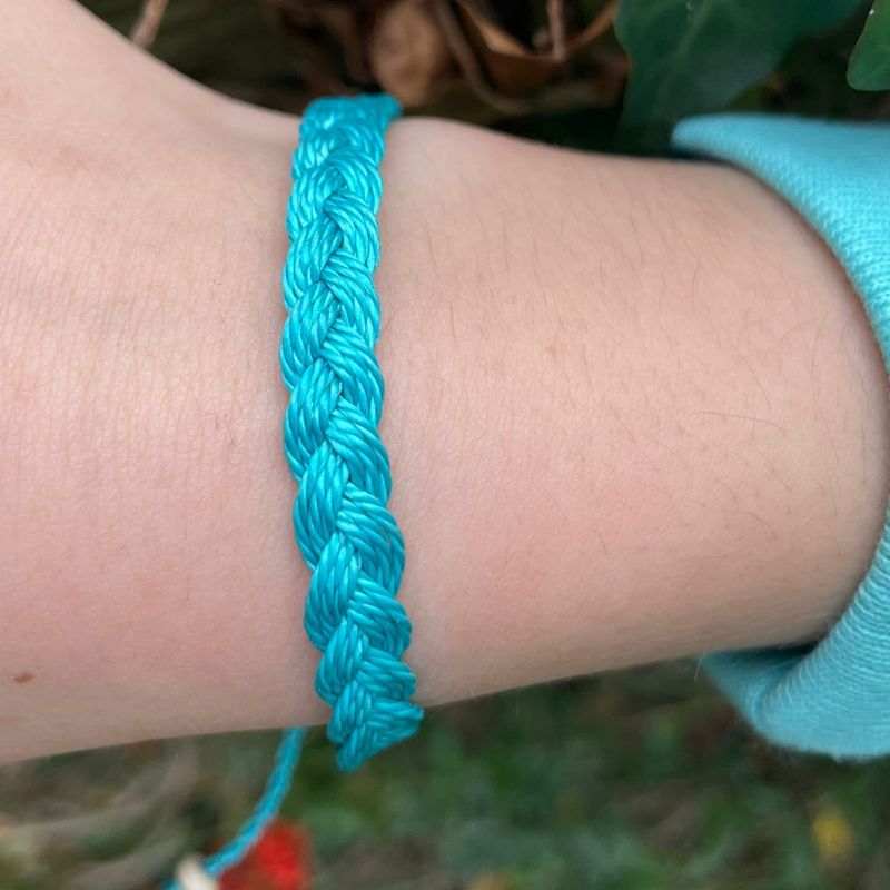 Close-up of a person's wrist wearing a blue braided bracelet with a blurred natural background.